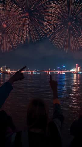 Vertical video: friends filming fireworks over riverbank at night, pointing and cheering