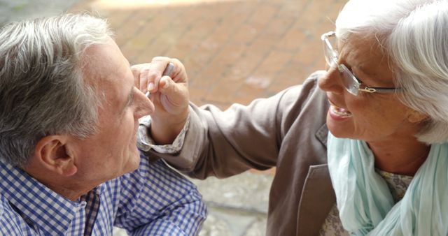 A senior Caucasian woman playfully touches the nose of a senior Caucasian man, both sharing a moment of joy and affection. Their laughter and close interaction suggest a deep bond, reflecting a long-term relationship or friendship.