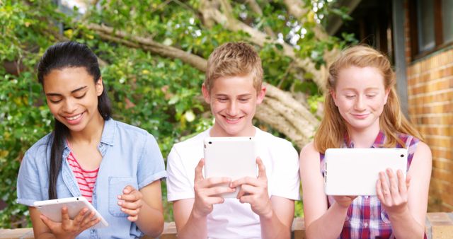 Happy teenagers sitting outdoors, using tablets. Image exudes positive emotions, friendship, and enjoyment. Ideal for articles on youth, technology in education, social bonding, and outdoor activities for kids.