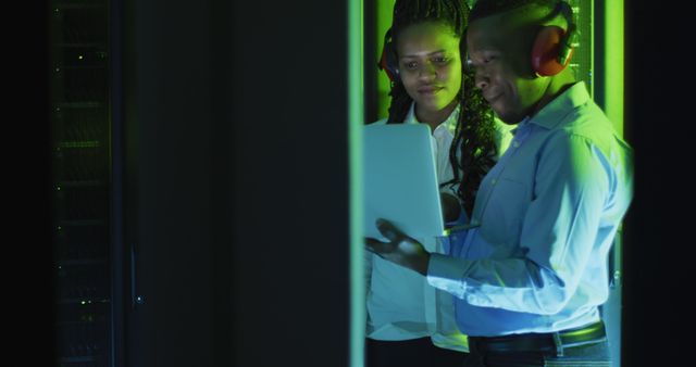 Two engineers are optimizing servers in a data center using a laptop. They are wearing headphones, focusing on their work in the green-lit server room. This can be used to depict professionals in the information technology field, data management, teamwork in high-tech settings, and IT infrastructure operations.