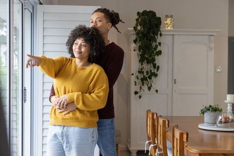Couple embracing by sliding glass doors, woman pointing, cozy sunlit home interior
