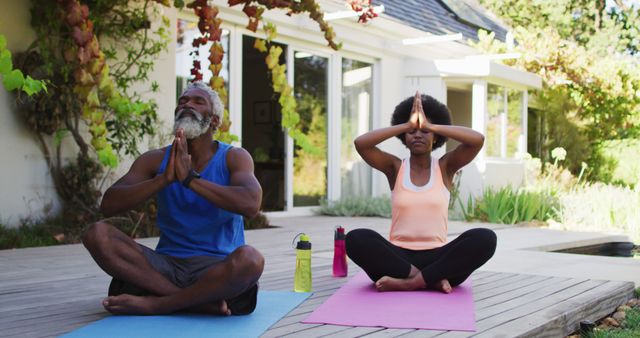 Senior couple meditating on yoga mats outdoors by their home. They are focusing on relaxation and exercise, enjoying a healthy lifestyle together. A great image for promoting senior fitness, wellness programs, yoga classes, and activities for older adults in a natural setting.