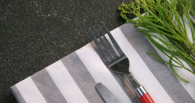 A fork and knife rest on a striped napkin beside fresh dill on a dark surface, with copy space. It suggests a setting prepared for a meal, highlighting the importance of fresh ingredients in culinary presentations.