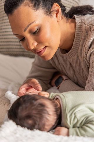 Mother leaning over newborn soothing baby on bed wearing smartwatch intimate nurturing