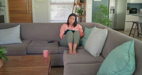 Mature woman relaxing on gray sectional sofa writing in notebook in cozy modern living room