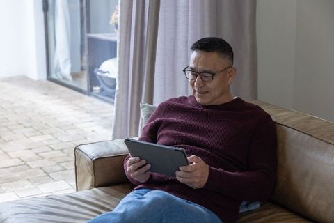 Senior man relaxing on sofa using tablet by glass door