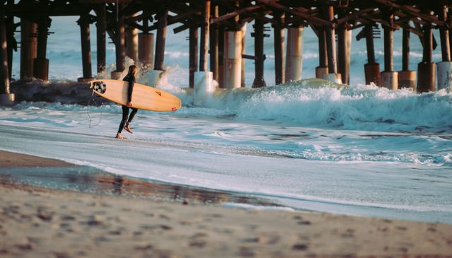 Surfer stands near ocean holding surfboard and looking at waves under wooden pier during sunset. Image conveys sense of adventure and outdoor lifestyle, ideal for promoting beaches, travel destinations, adventure sports, and active lifestyles.