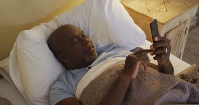 Mature man relaxing in bed while browsing his smartphone, showcasing modern technology and casual lifestyle. Ideal for use in content about technology adoption among seniors, relaxation, lifestyle habits, or nighttime routines.