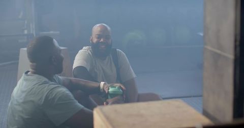 Male friends sitting and sharing green shaker during gym rest, laughing and chatting