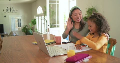 Smiling mother tutoring daughter during homeschooling at sunny dining table with laptop