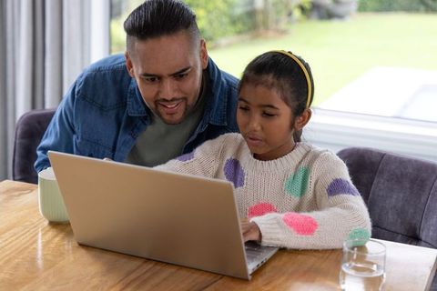 Father helping daughter with laptop at dining table learning together at home