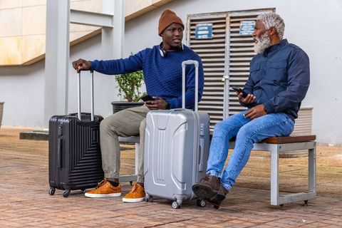 Travelers waiting and chatting on bench with suitcases and smartphones in urban plaza