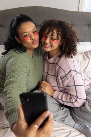 Mother and teen daughter taking selfie while wearing under-eye gel patches on cozy bed