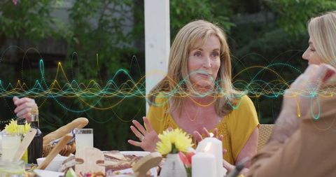 Mature woman talking and gesturing at outdoor dining table with audio waveform overlay