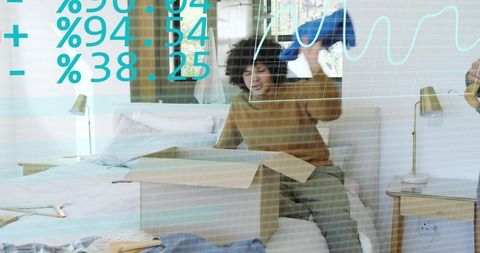 Young man unpacking clothes on bed, lifting blue jacket from box, casual home move