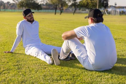 Cricket teammates sitting on sunlit grass wearing white uniforms and black caps talking