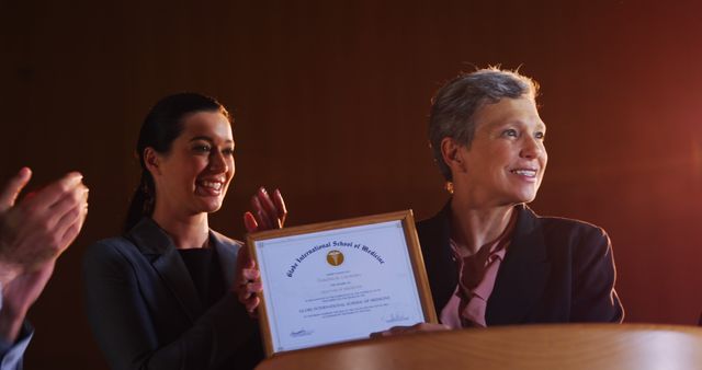 The image shows two businesswomen receiving an achievement award at a formal ceremony. The woman on the right is holding the framed certificate while the other claps. Great for use in business success stories, corporate events, award nominations, and professional achievements content.