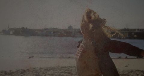 Backlit silhouette of woman standing with windblown hair on sandy beach near harbor pier