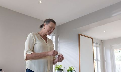 Senior woman standing in bright living room examining small object near leaning mirror