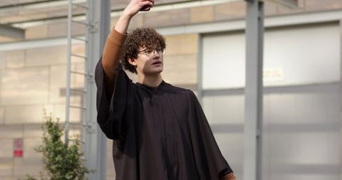 Graduate raising arm holding smartphone wearing black academic gown on urban campus