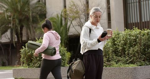 Mature woman checking smartphone while holding coffee, yoga mat-carrying woman walking urban plaza