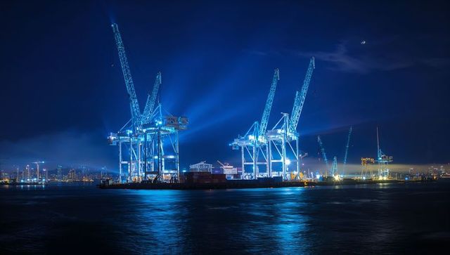 Blue-lit container cranes illuminating night harbor with reflections on calm water