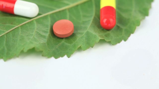 Image shows red and yellow pills placed on vibrant green leaf, symbolizing the intersection of modern medicine with nature. Useful for illustrating natural alternatives to pharmaceuticals or blending traditional botanical remedies with contemporary healthcare approaches in medicine-oriented publications or health blogs.