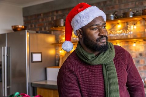 African american man wearing santa hat and green scarf standing in cozy rustic kitchen