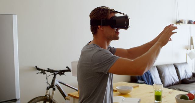 A young man in casual attire is experiencing virtual reality in a modern living room. He is wearing a VR headset and interacting with an unseen digital environment. The setting includes a bicycle, furniture, and natural light. This image is ideal for illustrating themes related to modern technology, virtual reality advancements, home entertainment, and immersive experiences.