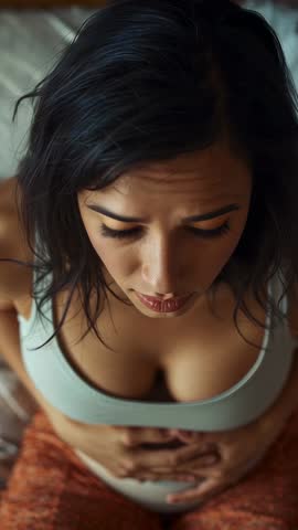 Vertical video: woman checking abdomen, holding stomach and looking up from bed, contemplative self-