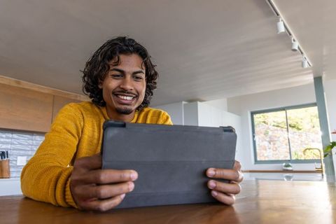 Indian man smiling while holding tablet at sunlit kitchen table
