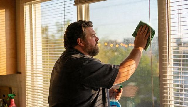 Man cleaning window with green microfiber cloth and spray bottle in sunlit home