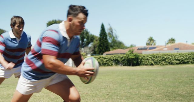 Intense rugby players engage in a dynamic practice session on an outdoor field. With focus and determination, one player is seen gripping the rugby ball tightly, showcasing action and agility. Ideal for use in sports promotions, fitness content, and articles highlighting team sports or physical training.