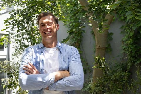 Smiling man standing with arms crossed in leafy backyard, casual summer lifestyle portrait