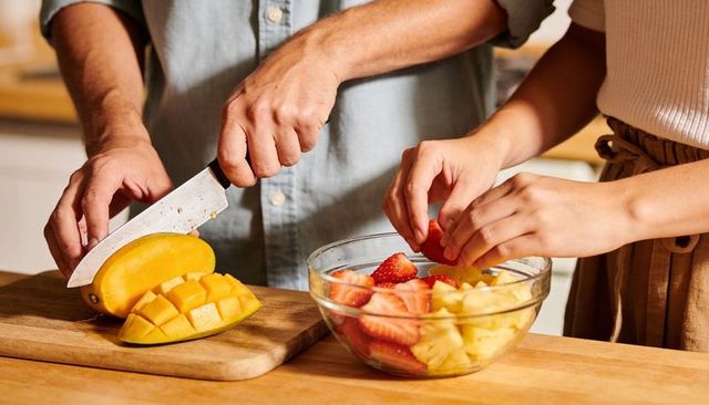 Couple preparing fresh fruit bowl, slicing mango and strawberries on wooden counter