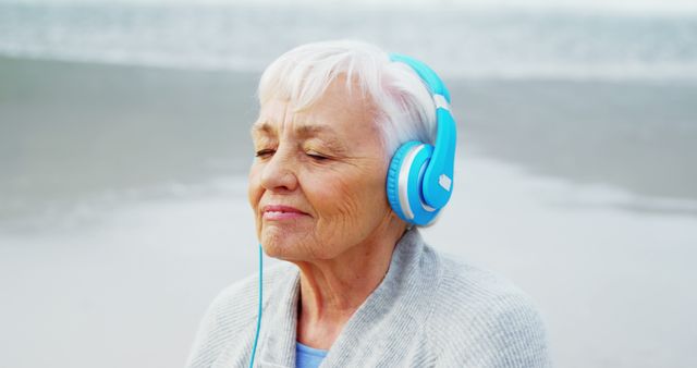A senior Caucasian woman enjoys music on her blue headphones, with copy space. Her peaceful expression suggests a moment of relaxation and contentment by the sea.