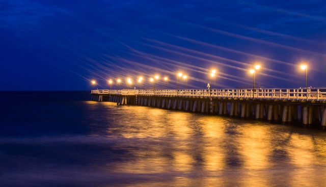 Pier with lights reflecting in water during nighttime, creating peaceful and tranquil scene. Suitable for travel brochures, waterfront property ads, relaxation-themed content, and background for night-related themes. Captures calm evening ambiance and serene coastal environment.