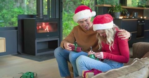Diverse couple in santa hats tagging holiday gifts by fireside in modern living room