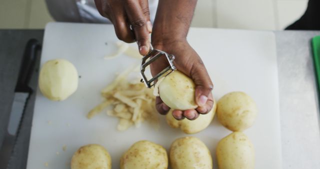 Chef peeling potatoes on cutting board with peeler in professional kitchen. Useful for illustrating cooking techniques, food preparation tutorials, restaurant kitchens, or kitchen tool demonstrations.