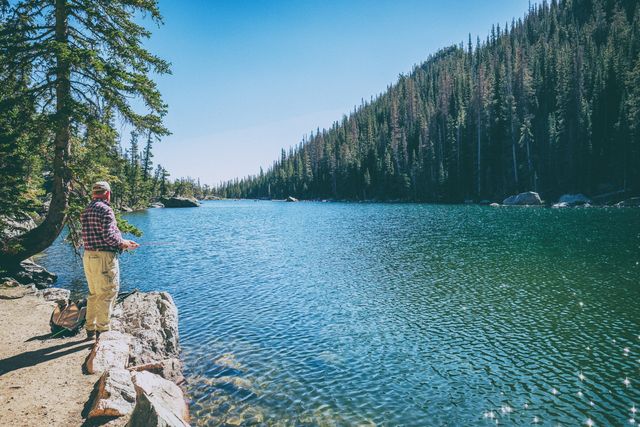 Senior man enjoying fishing at a picturesque mountain lake, surrounded by dense forest and calm waters. Ideal for promoting outdoor activities, retirement hobbies, travel destinations, and nature retreats.