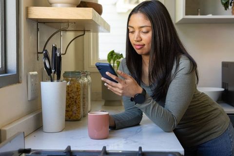 Asian woman leaning on modern kitchen counter checking smartphone while holding pink mug