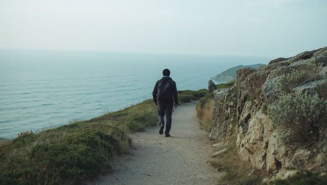 Walking solo hiker following coastal cliff trail with backpack overlooking calm sea