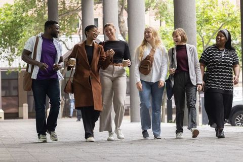 Multicultural college friends walking on campus colonnade carrying coffee and laughing
