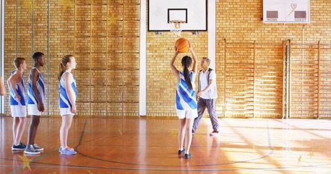 Teen basketball team practicing free-throw drill in sunlit school gym with coach guidance