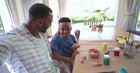 African american father and son painting colorful eggs at sunlit dining table