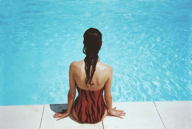 Woman in a red swimsuit with black patterns sits at the edge of a swimming pool, wet hair cascading down her back. Bright blue water and sun suggest a relaxing summer day. Ideal for travel, health and wellness promotions. Use for advertising swimwear, vacation spots, or lifestyle blogs.