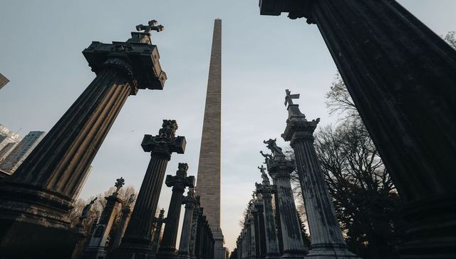 Obelisk rising through weathered memorial columns along symmetrical urban corridor at dusk
