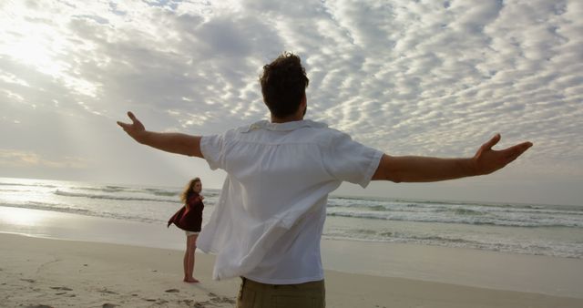 Man with open arms appreciating ocean breeze with female partner nearby on calm sandy beach. Ideal for beach vacation promotions, travel blogs, romantic getaway ads, and lifestyle content.