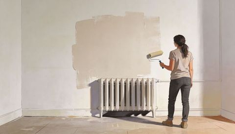 Woman Painting Wall with Roller Over Radiator in Bright Empty Room Neutral Beige Renovation