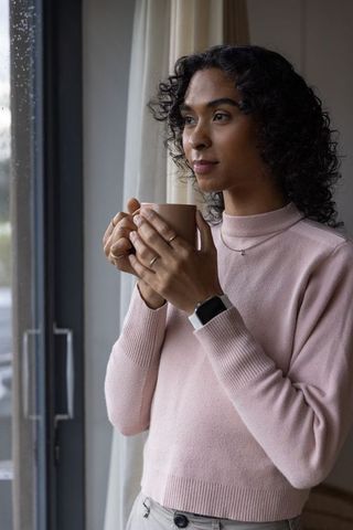 Nonbinary adult holding mug by rainy window, gazing contemplatively in pink sweater